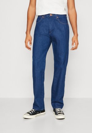 Mann trägt dunkelblaue Jeans mit geradem Bein und schwarze Low-Top-Sneakers mit weißen Schnürsenkeln, steht vor einem einfarbigen Hintergrund.