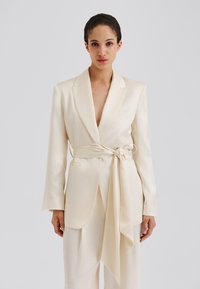 Krémovo sfarbený saténový blazer s dlhými rukávmi, hlbokým výstrihom do V a opaskom viazaným v páse. Má dve predné vrecká a hladkú, luxusnú textúru.