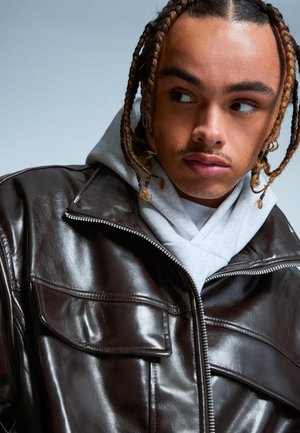 Brown leather jacket with a front zipper, chest pockets, and a soft gray hoodie underneath. Smooth texture and structured design.