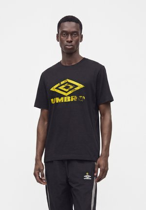 Άνδρας με μαύρο μπλουζάκι Umbro με φθαρμένο κίτρινο λογότυπο και μαύρη φόρμα με λευκές πλευρικές ρίγες και λογότυπο Umbro.