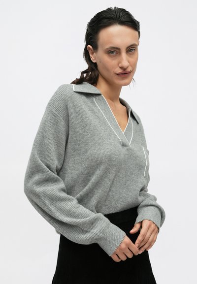Grauer Strickpullover mit V-Ausschnitt und Kragen, der weiße Paspeln an den Rändern und gerippte Bündchen aufweist. Das Modell trägt ihn über einem schwarzen Rock.