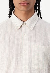 Chemise à boutons beige clair en tissu texturé et froissé. Présente un motif à carreaux, une poche poitrine et un col classique.