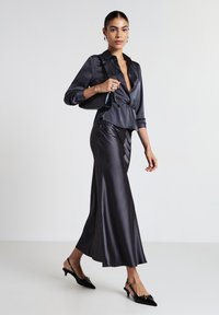 Blusa di raso scuro con un nastro in vita, abbinata a una lunga gonna di raso. Tacchi slingback neri con punta affilata e accenti dorati.