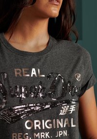 Camiseta de algodón gris con un logo de lentejuelas en plata y negro, de manga corta y cuello redondo, que presenta texto y acentos de la marca.