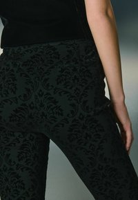 Pantaloni negri din catifea, cu un model texturat de damasc. Viziunea din spate pune în evidență buzunarele și o siluetă ajustată.