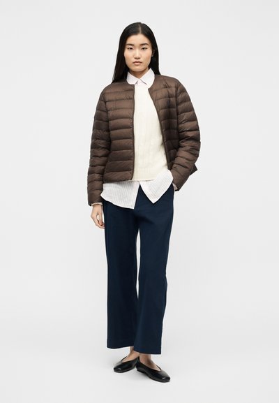 Γυναίκα όρθια που φοράει καφέ μπουφάν με καπιτονέ ύφασμα, εκρού πλεκτό πουλόβερ με κορδόνια πάνω από ριγέ πουκάμισο, μπλε σκούρο παντελόνι με φαρδιά μπατζάκια και μαύρες μπαλαρίνες.