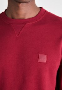 Sweat-shirt bordeaux avec un col rond, présentant un petit patch carré avec l'inscription "BOSS" sur la poitrine. Tissu doux et lisse.