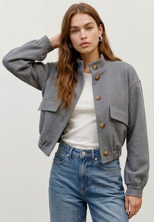 Jeune femme aux longs cheveux ondulés portant des boucles d'oreilles créoles argentées, une veste grise boutonnée, une chemise blanche et un jean bleu taille haute.