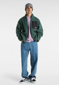 Grön fleecejacka med en svart dragkedjorficka, i kombination med en ljuslila t-shirt och ljusblå baggyjeans. Svarta sneakers med vita snören.