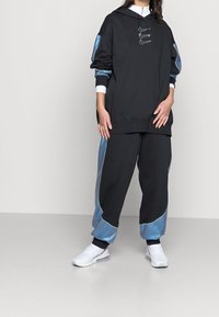 Svart huvtröja med blå satängdetaljer och vita muddar, tillsammans med svarta sweatpants med blå sidpaneler. Vita sneakers fulländar outfiten.