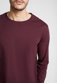 Bordeauxfarbenes Langarmhemd aus weichem, glattem Stoff. Verfügt über einen runden Halsausschnitt und schmale Ärmel. Minimalistisches Design, ohne Muster.