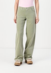 Pantalon en velours côtelé vert avec un design à jambes larges, comportant des poches avant et une fermeture par bouton. Assorti avec des chaussures beiges.