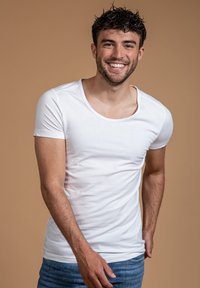 Witte T-shirt met korte mouwen en een rond decolleté, gemaakt van een gladde stof, gedragen met blauwe jeans tegen een neutrale achtergrond.