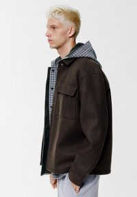 Giacca marrone oversize con tasca frontale, indossata sopra una camicia a quadri con cappuccio grigio. Tessuto liscio, vestibilità rilassata, accessori minimi.