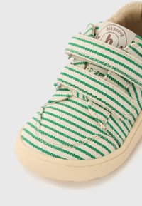 Πράσινο και λευκό ριγέ πάνινο παπούτσι με λουράκια βέλκρο και φυσική σόλα από καουτσούκ· διαθέτει κυκλικό λογότυπο στη γλώσσα του παπουτσιού.