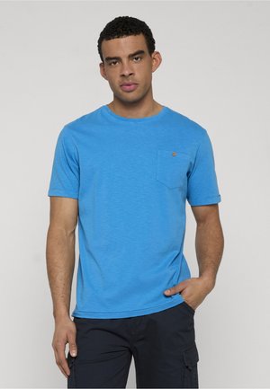 Mężczyzna z krótkimi kręconymi włosami, ubrany w jasnoniebieską koszulę z krótkim rękawem i ciemne spodnie cargo, stoi z jedną ręką w kieszeni.