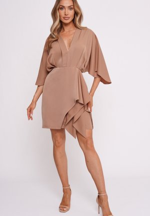 Woman wearing a beige wrap-style short dress with wide sleeves, a deep V-neck, and ruffle detail, paired with nude high-heeled sandals.