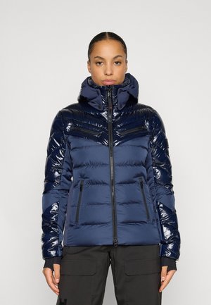 Kobieta ubrana w granatową, pikowaną zimową kurtkę z błyszczącymi panelami, z zamkiem z przodu, dwoma kieszeniami na piersi i dwoma kieszeniami bocznymi na zamek, stoi na jednolitym tle.