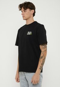 Joven con tatuajes en el brazo derecho, lleva una camiseta negra de manga corta con texto blanco en el pecho y pantalones gris claro, de pie de lado.