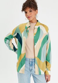 Chemise colorée à boutons avec des motifs ondulés verts, bleus et beiges, en tissu léger. Assortie avec un haut neutre et un jeans bleu.