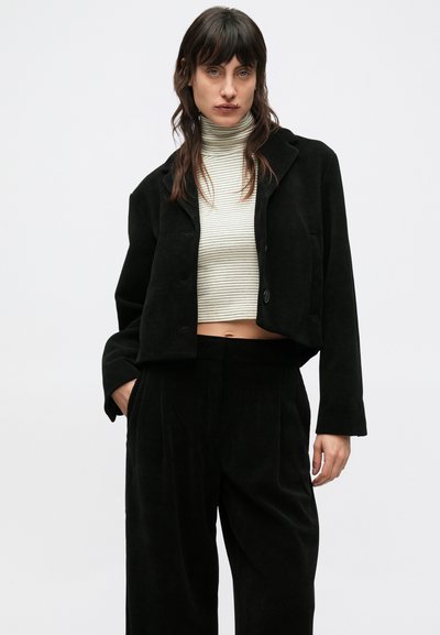 Fekete kordbársony rövidített zakó galléros nyakkivágással, gombokkal és elől zsebekkel, hozzáillő fekete nadrággal párosítva.