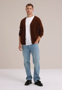 Bruine gebreide cardigan met knopen, gedragen over een wit T-shirt, gecombineerd met lichtblauwe jeans en zwarte schoenen tegen een neutrale achtergrond.