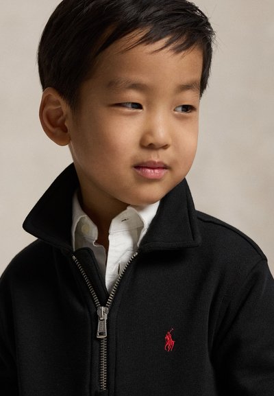 Jeune garçon aux cheveux noirs courts, portant un pull noir zippé par-dessus une chemise blanche à col, regardant légèrement sur le côté.