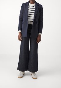 Blazer navy sopra a un maglione a righe bianche in maglia, abbinato a jeans in denim scuro a gamba larga e sneaker chiare. Morbidezza dei tessuti e design a strati.