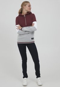 Haut à capuche zippé bordeaux et gris avec des manches blanches, doté d'une fermeture à glissière 1/4, d'une bande contrastante à l'ourlet et d'un patch logo sur le côté inférieur. Associé à un jean foncé et des baskets blanches.