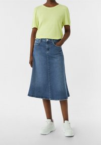 Blusa verde claro de manga corta combinada con una falda de mezclilla azul que llega a la rodilla, con bolsillos y un ligero corte en A, llevada con zapatillas blancas.