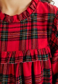Vestido de cuadros rojos con escote volado y dobladillo en capas con volantes. El tejido tiene una textura suave con acentos en verde, amarillo y negro en el patrón.