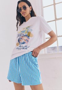 T-shirt bianco con una grafica di frutti di mare colorata e testo "Il pesce è fresco", abbinato a pantaloni corti a righe blu e bianche.