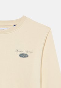 Μπεζ φούτερ με στρογγυλή λαιμόκοψη και το κείμενο «Denim Attitude», με ένα μικρό οβάλ λογότυπο που αναγράφει «Superior Quality Jack & Jones» στο στήθος.