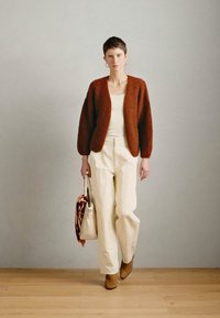 Rustfarvet fuzzy cardigan over hvid top, hvide vidde bukser og brune ankelstøvler. Modellen holder en mønstret taske. Trægulvsbaggrund.