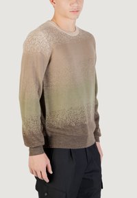 Pletený svetr s gradientním vzorem od béžové po zelenou, s texturovanými akcenty a žebrovaným límcem a manžetami. Model má na sobě černé kalhoty.
