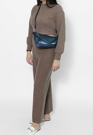 Femme en tenue de détente marron composée de deux pièces, portant un sac bandoulière bleu et des chaussures blanches sans lacets, debout devant un fond uni.