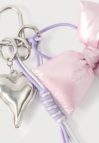 Μπρελόκ με ασημένιο γοητευτικό καρδιά, ροζ σατέν φιόγκο και λεβάντα κορδόνια με ασημένιες λεπτομέρειες, δημιουργώντας ένα στρωματοποιημένο, υφασμάτινο σχέδιο.