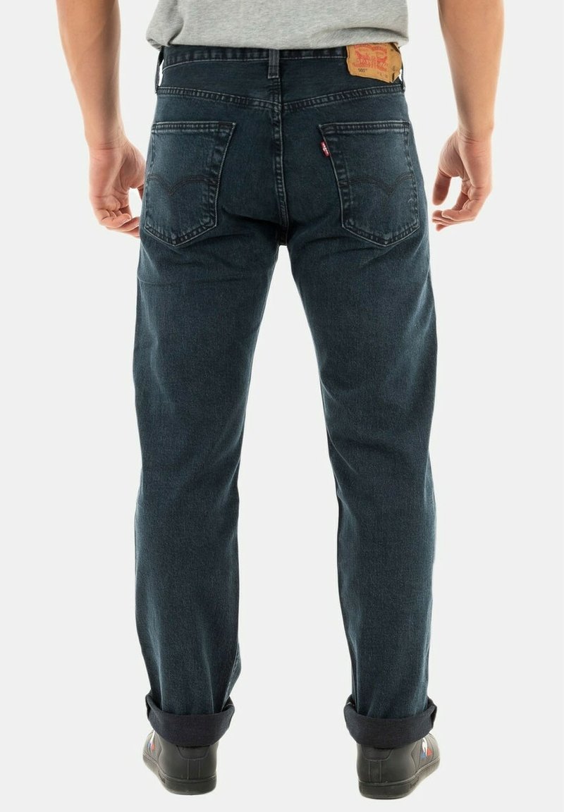 Hombre de pie con la espalda hacia la cámara, vistiendo jeans de mezclilla azul oscuro con dobladillos doblados y zapatillas negras.