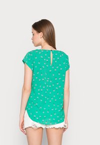 Blusa verde de mangas curtas com estampado floral, apresentando um detalhe em forma de chave na parte de trás, feita de um tecido leve e com uma textura suave.