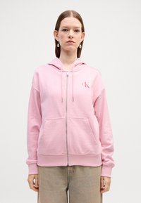 Felpa con zip di un delicato rosa chiaro, realizzata in tessuto morbido, dotata di una tasca frontale e di un piccolo logo bianco sul petto. Maniche lunghe con polsini.