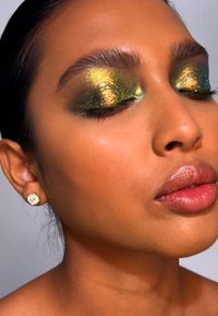 Nahaufnahme eines Gesichts mit kräftigem grünem und goldenem schimmerndem Lidschatten, glatter Haut und glänzenden nude Lippen. Kleine goldene Steckerohrringe werden getragen.