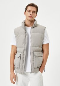 Chaleco acolchado gris claro con cuello alto, cierre frontal y dos bolsillos delanteros. Hecho de material acolchado, combinado con una camiseta blanca.