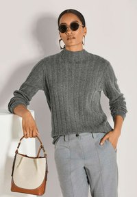 Grauer gerippter Strickpullover mit hohem Kragen, kombiniert mit hellgrauen, taillierten Hosen. Die Person hält eine beigefarbene und braune Handtasche. Runde schwarze Sonnenbrille.