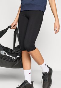 Μαύρο εφαρμοστό κολάν με μήκος μέχρι το γόνατο, φορεμένο με λευκές κάλτσες και μαύρα αθλητικά παπούτσια, κρατώντας μαύρη τσάντα duffel με λογότυπο.