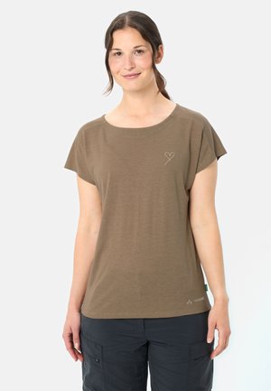 Frau trägt ein braunes kurzärmeliges T-Shirt mit kleinem Herzdesign und dunkle Cargohosen, steht vor einem schlichten hellen Hintergrund.