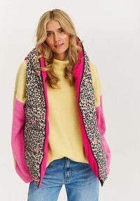 Pletená vesta s leopardím vzorem a jasně růžovými akcenty, přelivená přes žlutý svetr. Má vysoký límec a kapsy. Volný střih.