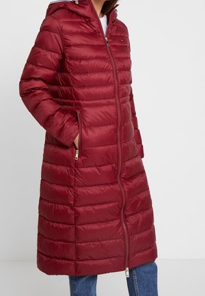 Femme portant un long manteau matelassé bordeaux avec fermeture éclair à l'avant et poches latérales, associé à un jean bleu.