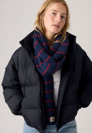Giovane donna con giacca a solo nero, sciarpa blu navy con strisce rosse, maglietta bianca e jeans azzurri chiari su uno sfondo neutro.