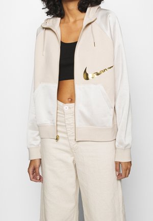 Femme portant un sweat à capuche zippé beige et blanc avec logo Nike doré, un crop top noir et un pantalon beige clair sur fond uni.