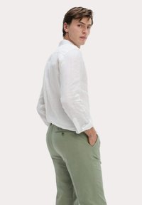Chemise blanche en lin à manches longues avec les poignets retroussés, assortie à un pantalon vert clair doté de poches. Le mannequin se tient debout avec une main dans une poche.